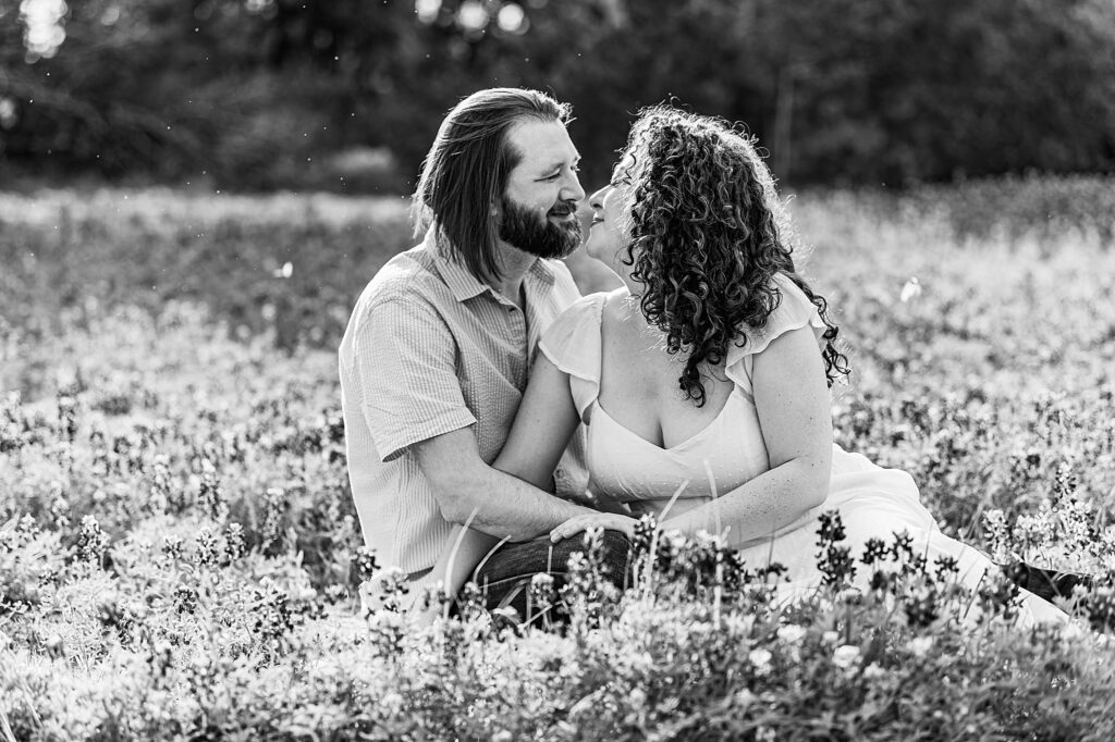 Black and White Photo of a couple sitting in a field of bluebonnets sharing a romantic moment. Dripping Springs Engagement Photographer Lydia Teague.
