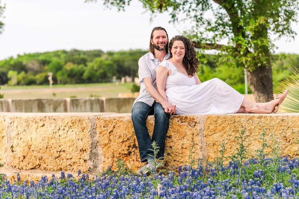 An engaged couple sitting on a limestone wall above a dense bed of blooming bluebonnets. Dripping Springs Engagement Photographer Lydia Teague
