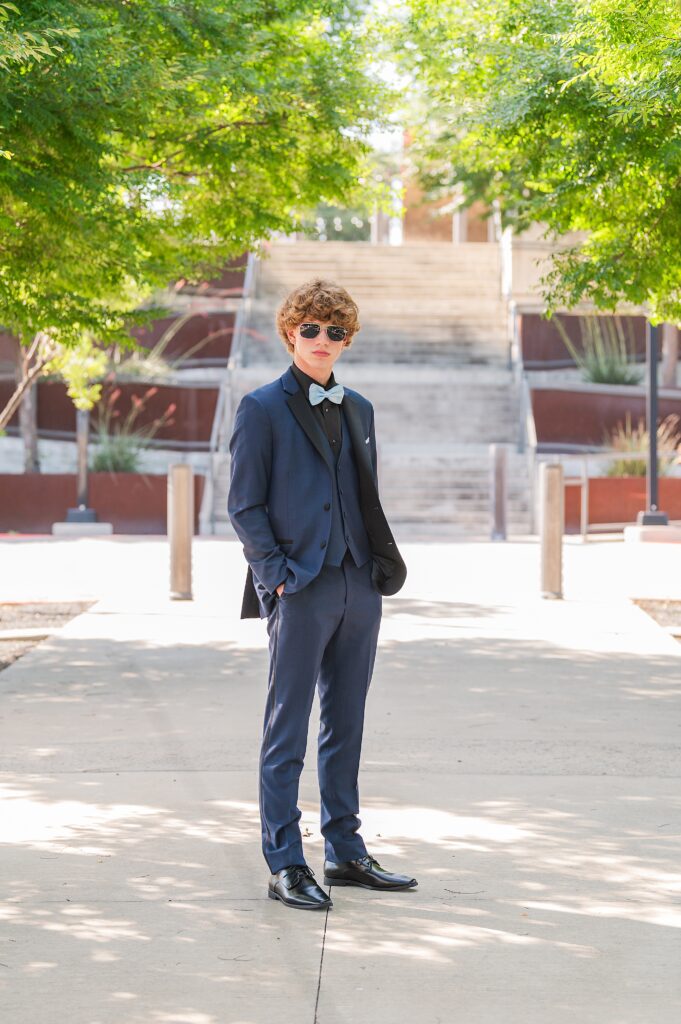 High School Junior wearing a Midnight Blue Tuxedo with light blue bow tie and pocket square. He's wearing sunglasses and posing on a tree lined sidewalk. Prom Portraits