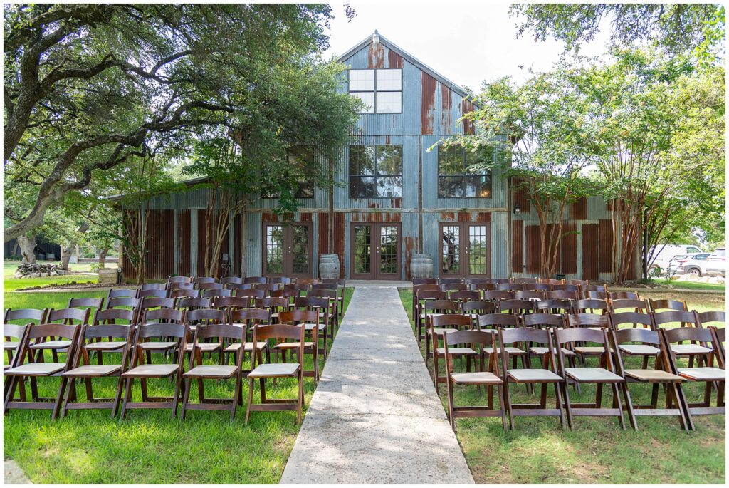 The rustic outdoor ceremony space at Vista West Ranch.  The building is covered with weathered and rusted tin. Folded wooden chairs with white seats are all set up in rows. 