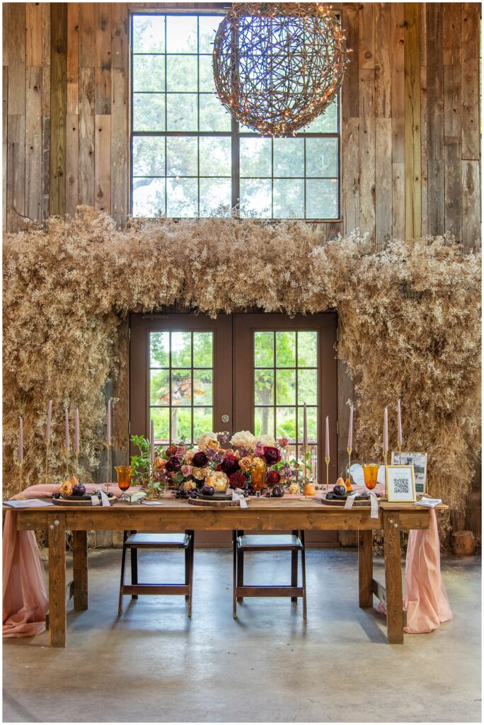 The inside of Vista West Ranch's Reception Barn.  Hanging grapevine balls with lights adorn the ceiling. Below is a table that is decorated with cream, red, and peach colored florals for a Wedding. 