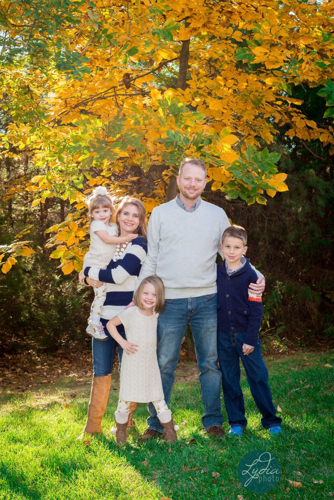 Family of 5 standing in front of a fall colored tree wearing cream and navy blue, photographed by Dripping Springs Texas based Lydia Teague Photography