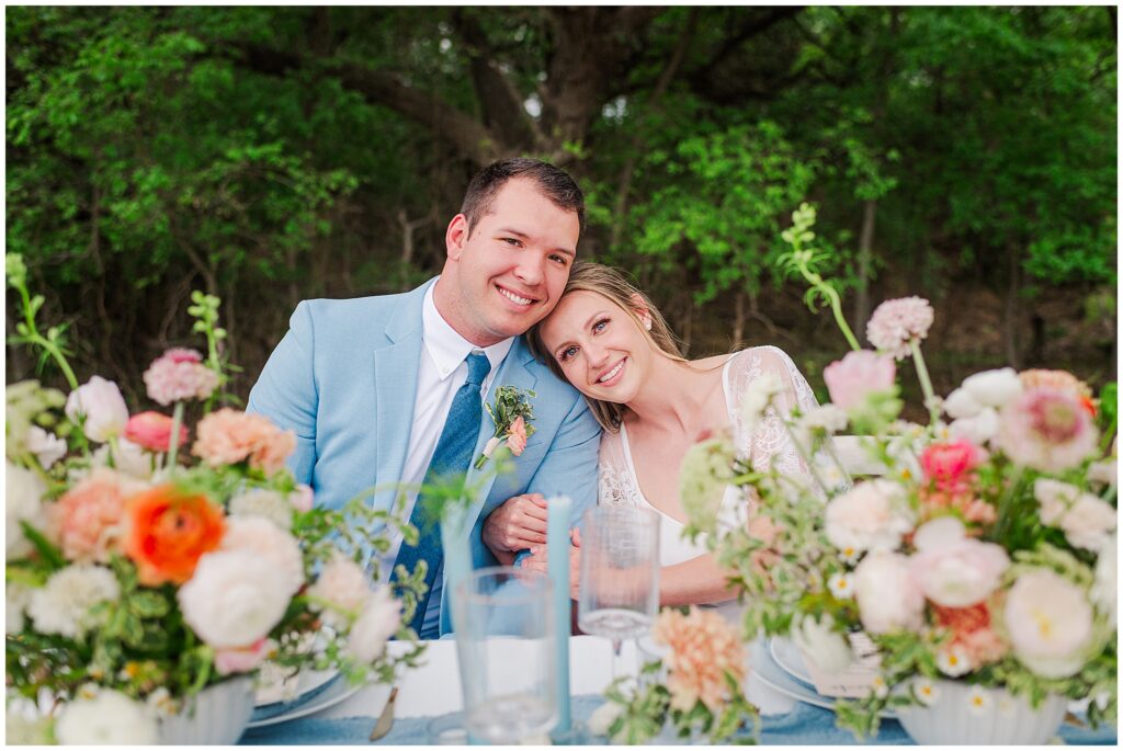 Groom is wearing a pastel blue jacket and sitting at the head table with his bride surrounded by pastel orange, peach, and pink flowers. Photo taken by Dripping Springs TX based Lydia Teague Photography. 