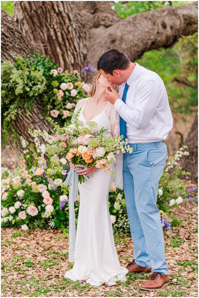 Bride and Groom kissing under large live oak trees in the Texas hill country surrounded by whimsical and wild flowers. Photo taken by Dripping Springs TX based Lydia Teague Photography. 